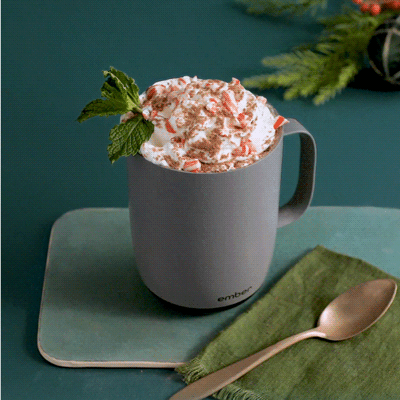 A gif of a hand placing a peppermint stick into the whipped cream that fills a Gray Ember Mug². The mug sits on a dark green background and there is a gold spoon laying ontop of a lighter green napkin in the foreground.