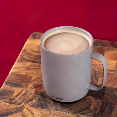 A gif of dark chocolate pouring over whip cream in a gray Ember Mug on a wooden cutting board with a red background.