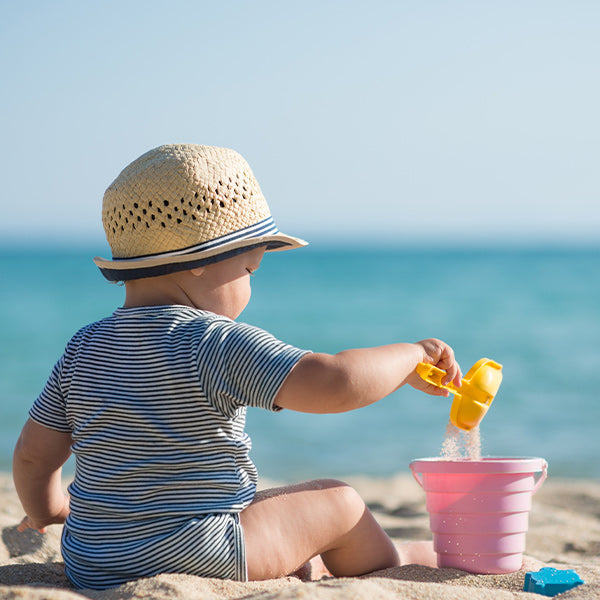 Baby in sunhat sits on beach playing with beach toys.