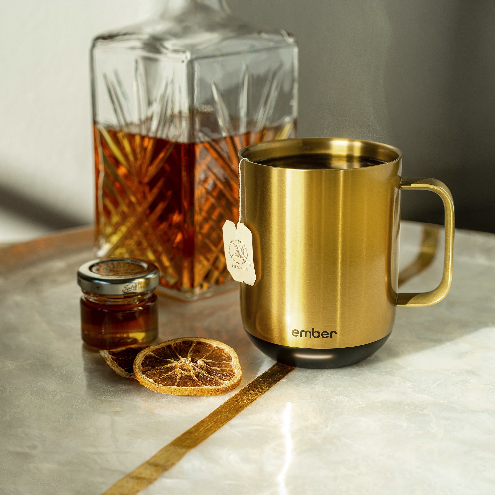 A Gold Ember Mug² filled with black tea sits on a marble and gold table next to a small jar of honey, slices of candied oranges, and a crystal carafe of a brown liquor.