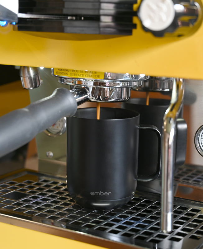 A black Ember Mug² sits underneath the spout of a chrome and yellow espresso machine.