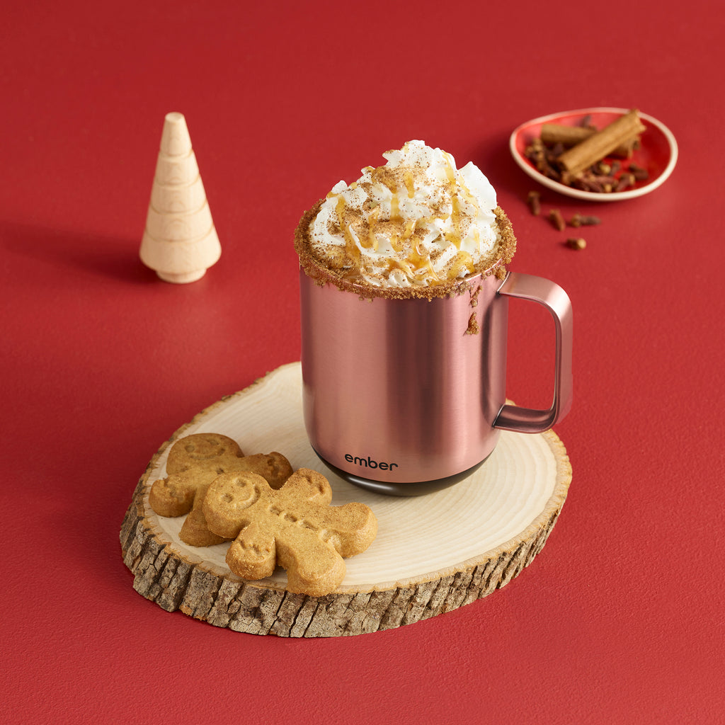 A Rose Gold Ember Mug² filled with whipped cream and cinnamon sits on a round cut of wood along wih two gingerbread cookies. The background is a deep red and is filled with gingerbread trees and cinnamon sticks.