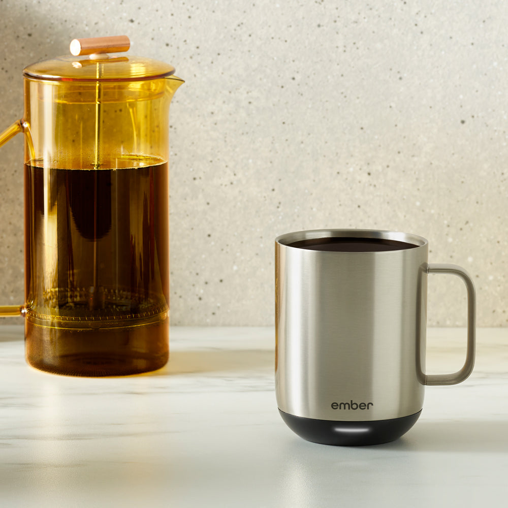 A stainless steel Ember Mug² sits next to a yellow french press filled with coffee.