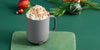 A Gray Ember Mug² is filled with whipped cream and topped with crushed candy cane and cinnamon. The mug sits on a dark green background filled with red and green holiday ornaments alongside a leafy branch with red berries.