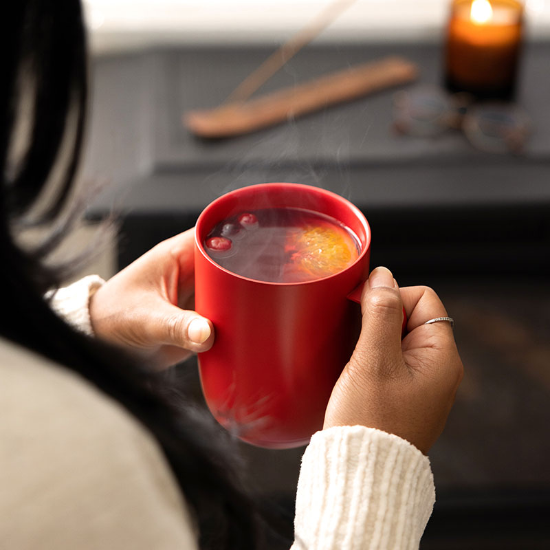 A Gray Ember Mug² is filled with a hot toddy drink and topped with apple slices. It sits on a weathered dark wooden table.