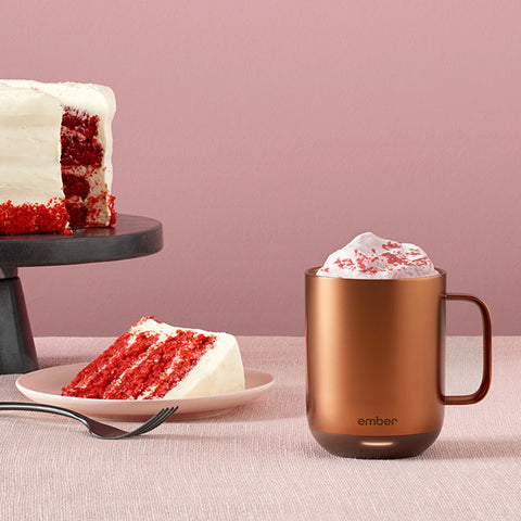 Ember Mug²: Copper Edition topped with whipped cream and red sprinkles sitting next to several slices of Red Velvet Cake on a pink background.